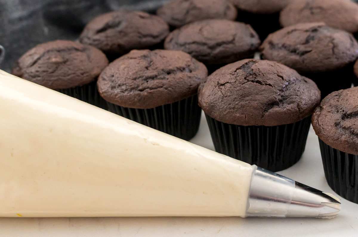 Closeup on a decorating bag filled with Buttercream Frosting sitting in front of a batch of unfrosted cupcakes.