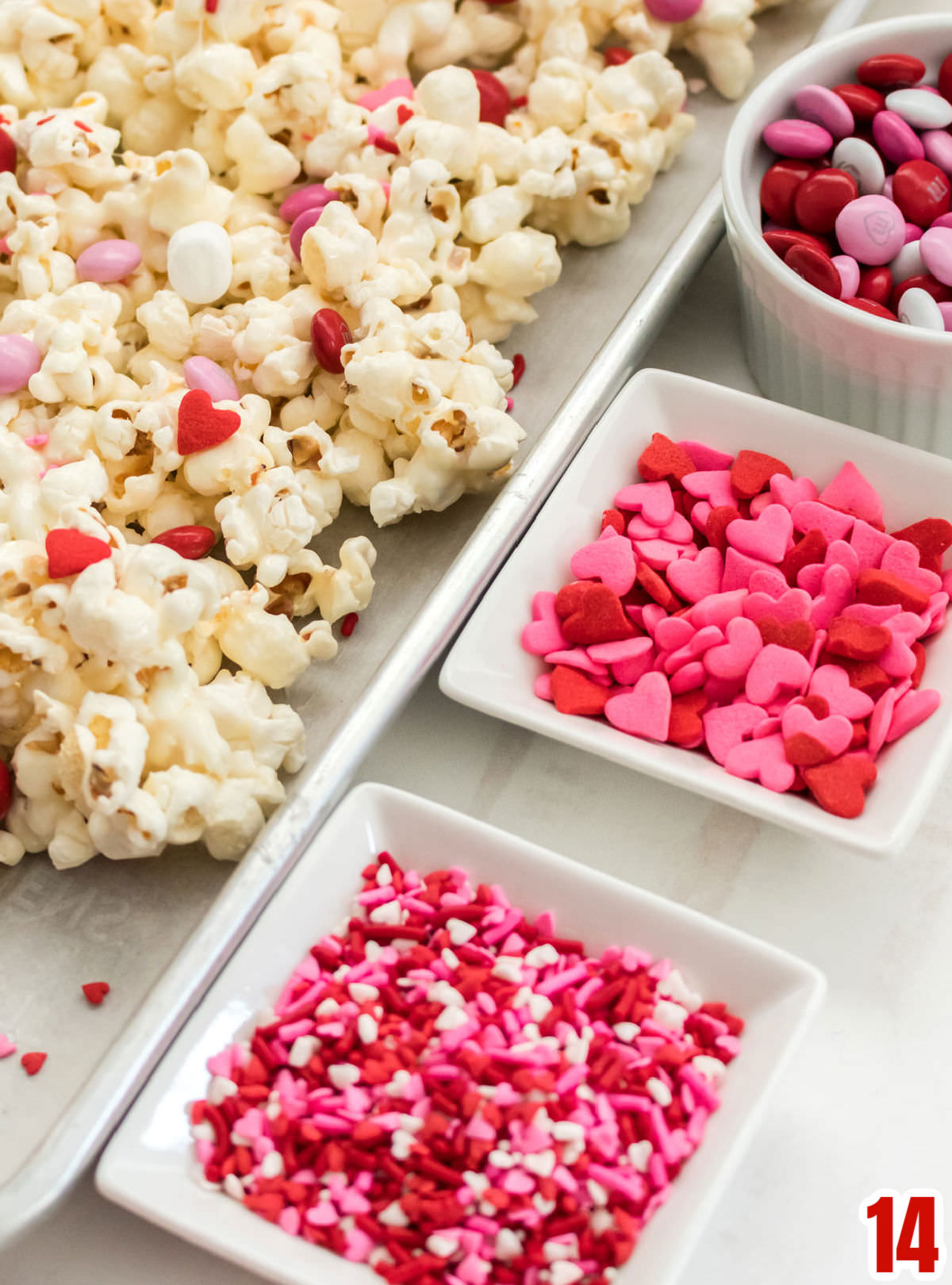 Close up of a three small white bowls filled with Valentine's sprinkles, Heart sprinkles and Valentine's Mix M&M's sitting next to a cookie sheet covered with Valentines  Day Popcorn.