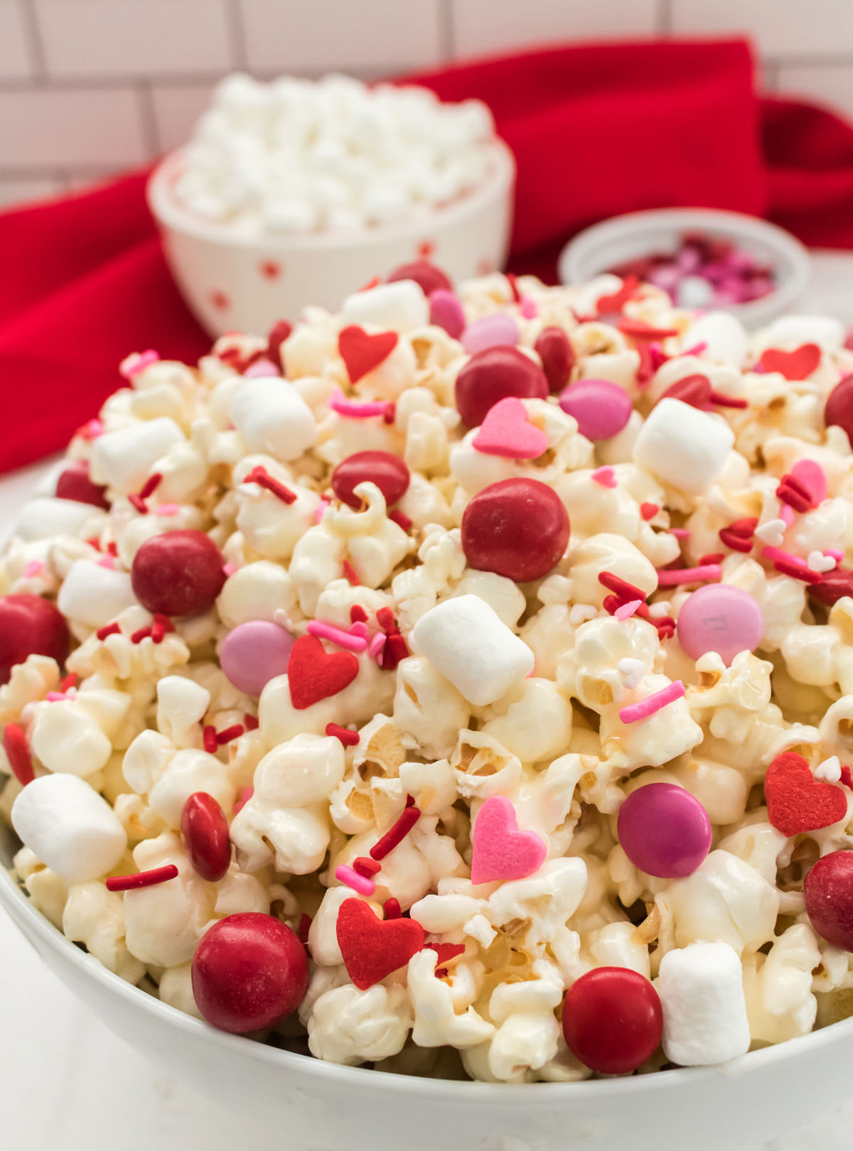 Close up of Valentine's Day Popcorn in a white bowl on a white table sitting in front of a bowl of marshmallows and M&M's.