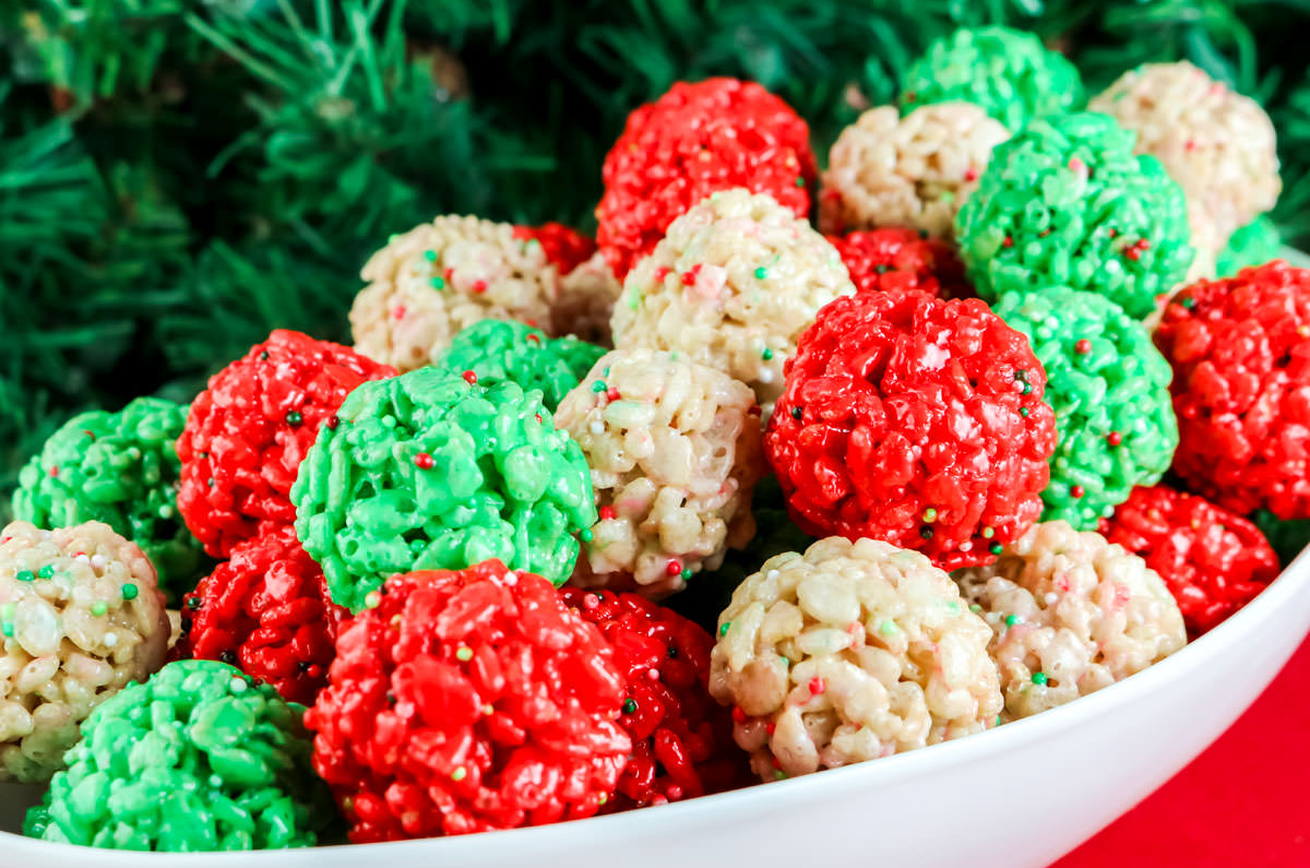 Closeup on a white serving bowl filled with Christmas Rice Krispie Treats that is sitting on a red table with Christmas greenery in the background.