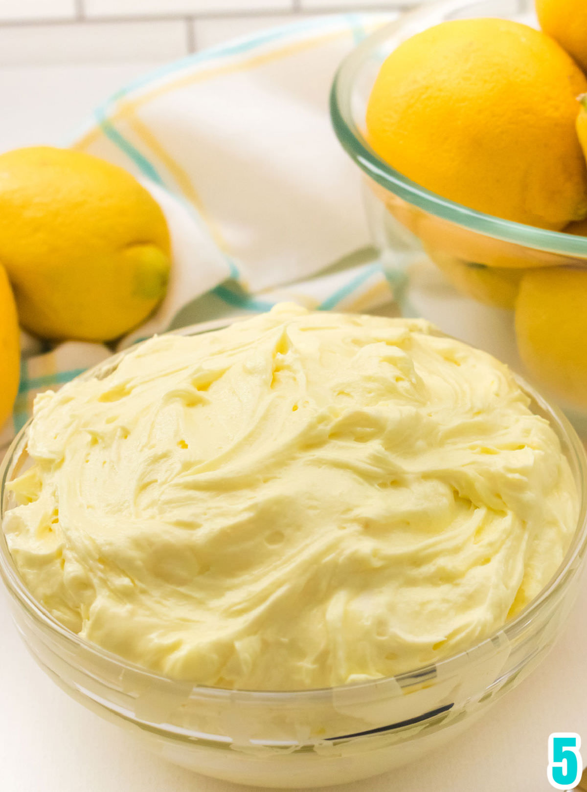 Closeup on a clear bowl filled with homemade Lemon Buttercream Frosting sitting in front of a bowl filled with fresh lemons.