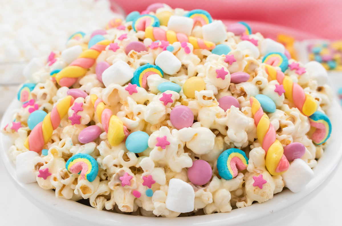 Closeup on a white serving bowl filled with Unicorn Popcorn with a ramekin of M&M's and a bowl of marshmallows in the background.