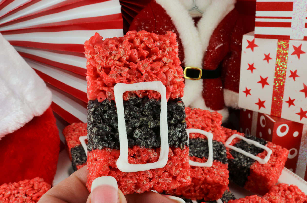 A hand holding up a single Santa Claus Rice Krispie Treat in front of a platter full of the rest of the batch.