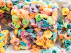 Close-up on a stack of two Froot Loops Marshmallow Treats sitting on a white table in front of glass bowls filled with Froot Loop Cereal and Mini Marshmallows.
